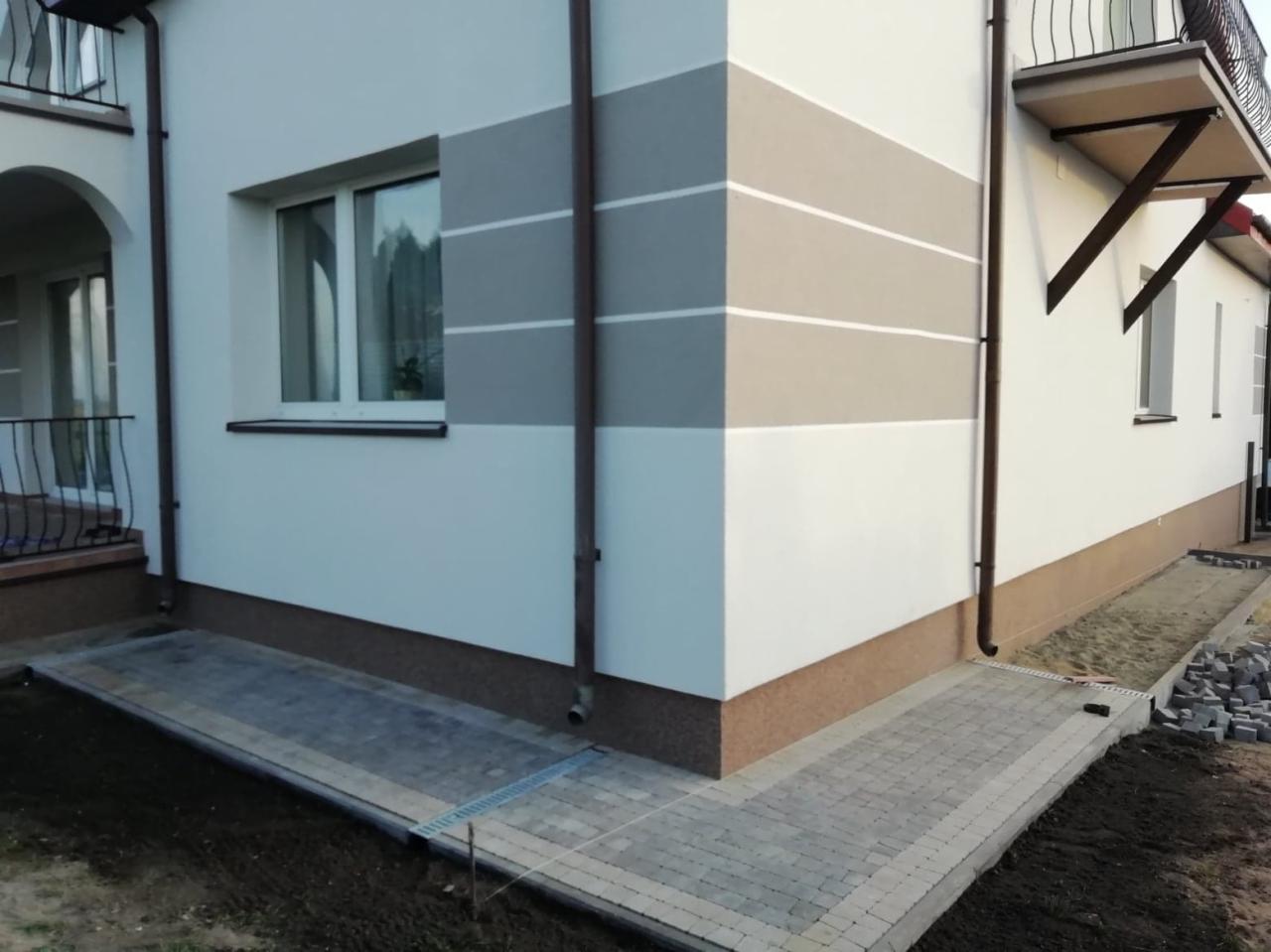 Dwukondygnacyjny budynek z ocieploną elewacją w odcieniach bieli i szarości, z brązowym cokołem i nową kostką brukową wokół, widoczne rury spustowe i okno z białymi ramami.