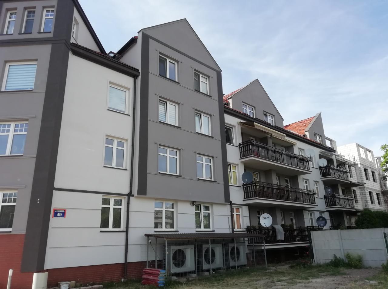 Odnowiony budynek wielorodzinny z szarą i białą elewacją, widoczne balkony z czarnymi balustradami i anteny satelitarne, numer budynku 49 na ścianie, oraz klimatyzatory na parterze.