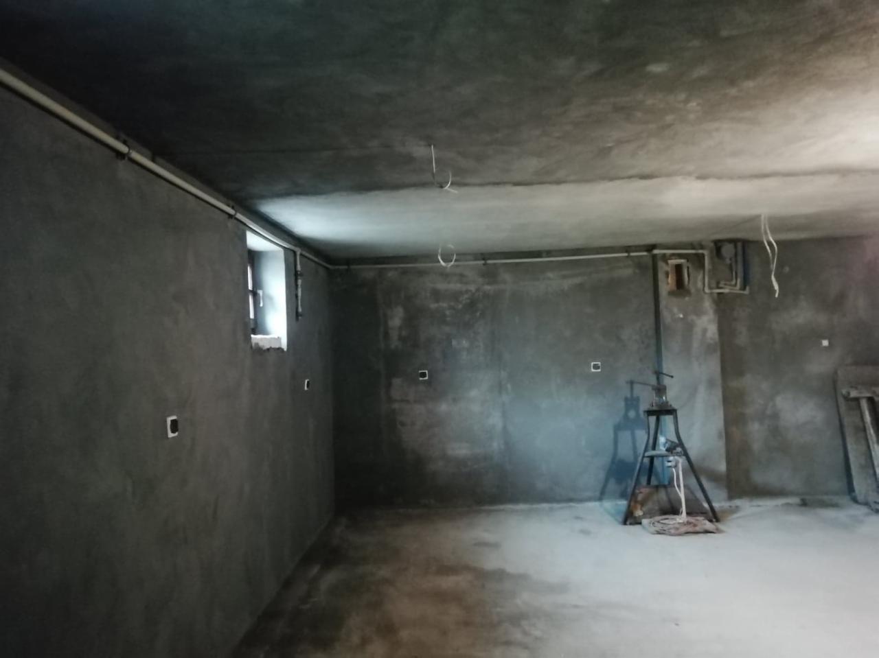 Surowe wnętrze w trakcie remontu: szare ściany z widoczną fakturą tynku, małe okno, instalacje elektryczne i hydrauliczne, podnośnik na trójnogu na środku pomieszczenia.