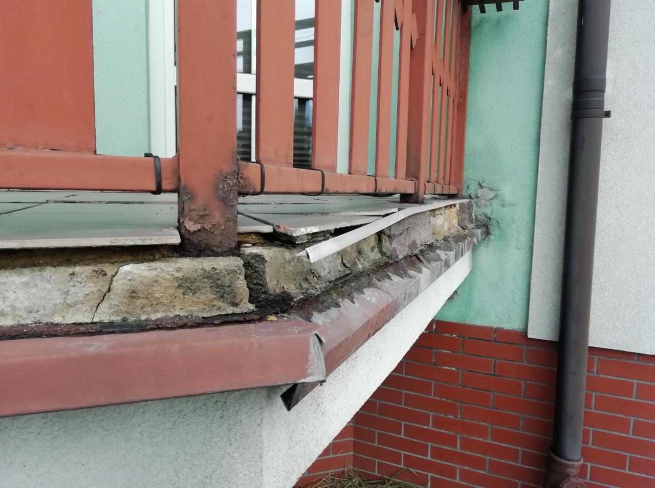 Zniszczony balkon z odpadającym tynkiem, skorodowaną metalową balustradą i uszkodzonymi płytkami podłogowymi.
