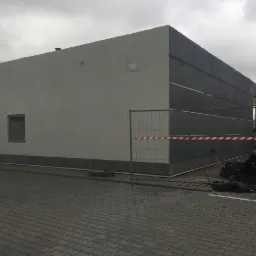 Dwukolorowa elewacja budynku komercyjnego w trakcie wykończenia, z widocznym ogrodzeniem tymczasowym i elementami rusztowania.