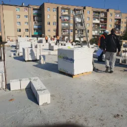 Budowa ściany z białych bloków silikatowych na fundamencie budynku wielorodzinnego, widoczni pracownicy w pomarańczowych kamizelkach i paleta z blokami marki Ekosilikat.