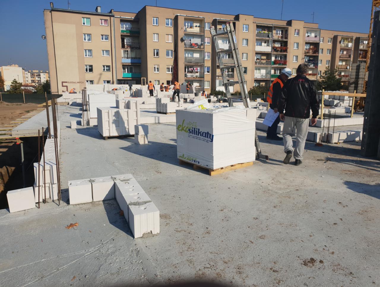 Budowa ściany z białych bloków silikatowych na fundamencie budynku wielorodzinnego, widoczni pracownicy w pomarańczowych kamizelkach i paleta z blokami marki Ekosilikat.