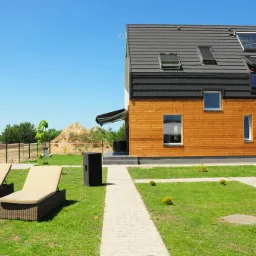 Nowoczesny dom jednorodzinny z drewnianą elewacją, solarem na dachu i leżakami w ogrodzie, widok z perspektywy trawnika w słoneczny dzień.