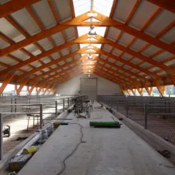 Wnętrze hali z drewnianą konstrukcją dachu, betonową podłogą i metalowymi barierkami, widok z perspektywy w głąb budynku.