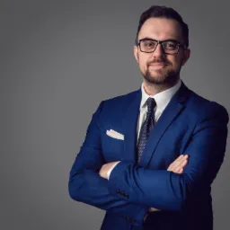 Portret uśmiechniętego adwokata w okularach, w niebieskim garniturze i z poszetką, na szarym tle.