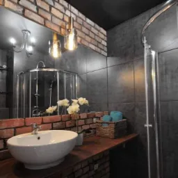 Nowoczesna łazienka z elementami cegły, ciemnymi płytkami i szklaną kabiną prysznicową typu walk-in, oświetlona wiszącymi lampami i z umywalką nablatową.