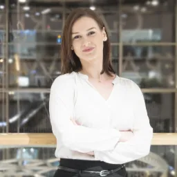 Portret kobiety z założonymi rękami, ubrana w białą bluzkę i czarną spódnicę, na rozmytym tle biurowym z elementami drewnianej balustrady.