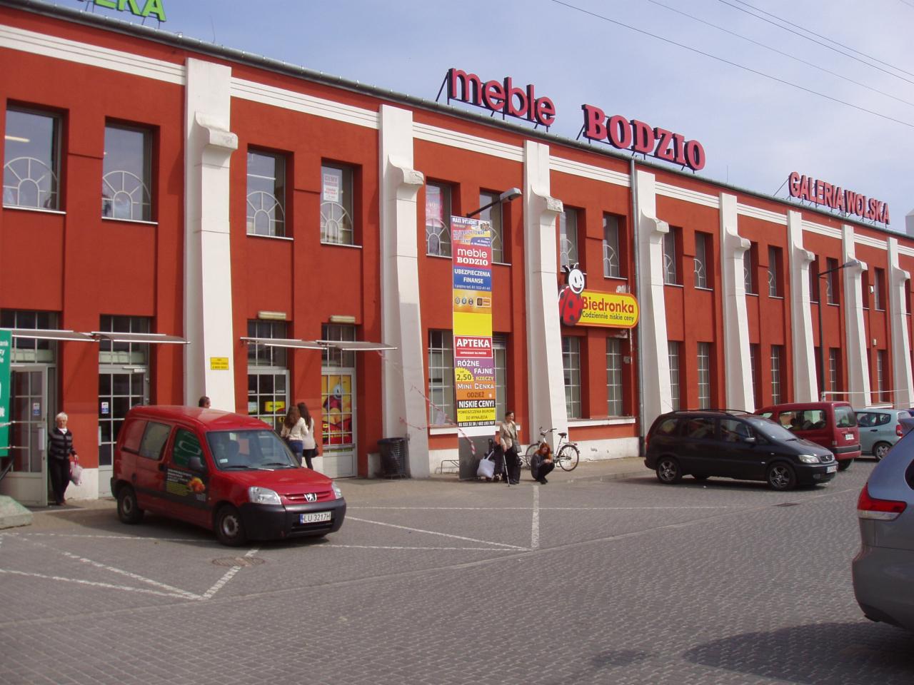 Czerwony budynek galerii handlowej z napisem 'meble BODZIO' nad wejściem, zaparkowane samochody i osoby przed wejściem.