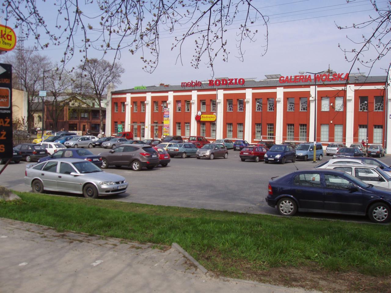 Widok na parking przed budynkiem z napisem 'meble RODZIO GALERIA WOLSKA', z widocznymi zaparkowanymi samochodami i drzewami z przodu