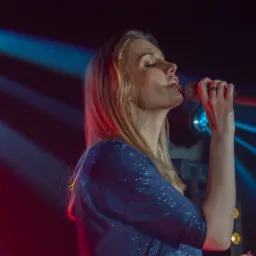 Wokalistka z blond włosami w błyszczącej, granatowej bluzce śpiewa do mikrofonu na scenie oświetlonej niebieskimi i czerwonymi światłami.