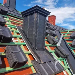 Montaż czarnej dachówki ceramicznej wokół komina wentylacyjnego na drewnianej więźbie dachowej pokrytej membraną, widok z góry na tle błękitnego nieba.