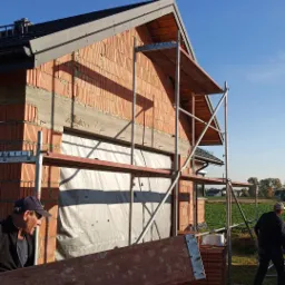 Budowa domu jednorodzinnego z czerwonej cegły, widoczne rusztowania, dach z ciemnej blachy, pracownik przenosi deskę, w tle pole i zabudowania.