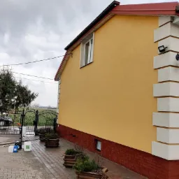 Żółta elewacja domu z ceglaną podmurówką, po umyciu, widoczny sprzęt do mycia ciśnieniowego na kostce brukowej, pochmurne niebo.