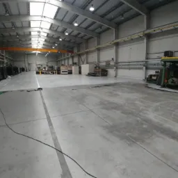 Przestronna hala przemysłowa z betonową posadzką, widoczne maszyny i urządzenia, oświetlenie sufitowe, suwnica pod stropem, ślady po czyszczeniu na podłodze, częściowo widoczny samochód z kablem...