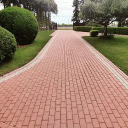 Czerwona kostka brukowa ułożona wzdłuż ścieżki, obramowana jaśniejszymi płytami, prowadząca przez zadbany ogród z równo przyciętymi krzewami i drzewami.