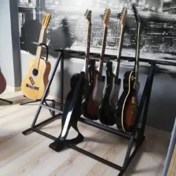 Czarny, metalowy stojak na pięć gitar różnych typów, z gitarą akustyczną umieszczoną obok, na tle fototapety z motywem miejskim.