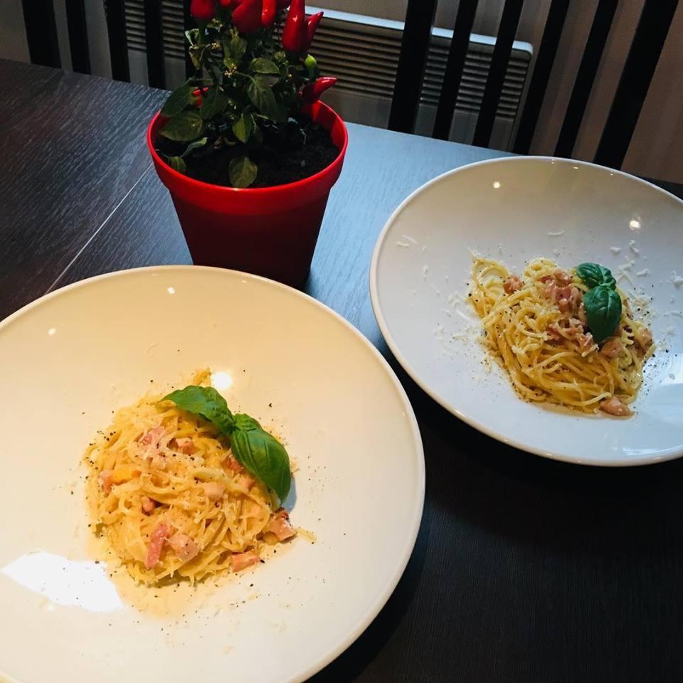 Dwa talerze spaghetti carbonara z boczkiem i bazylią na ciemnym stole, w tle doniczka z papryczkami chili.