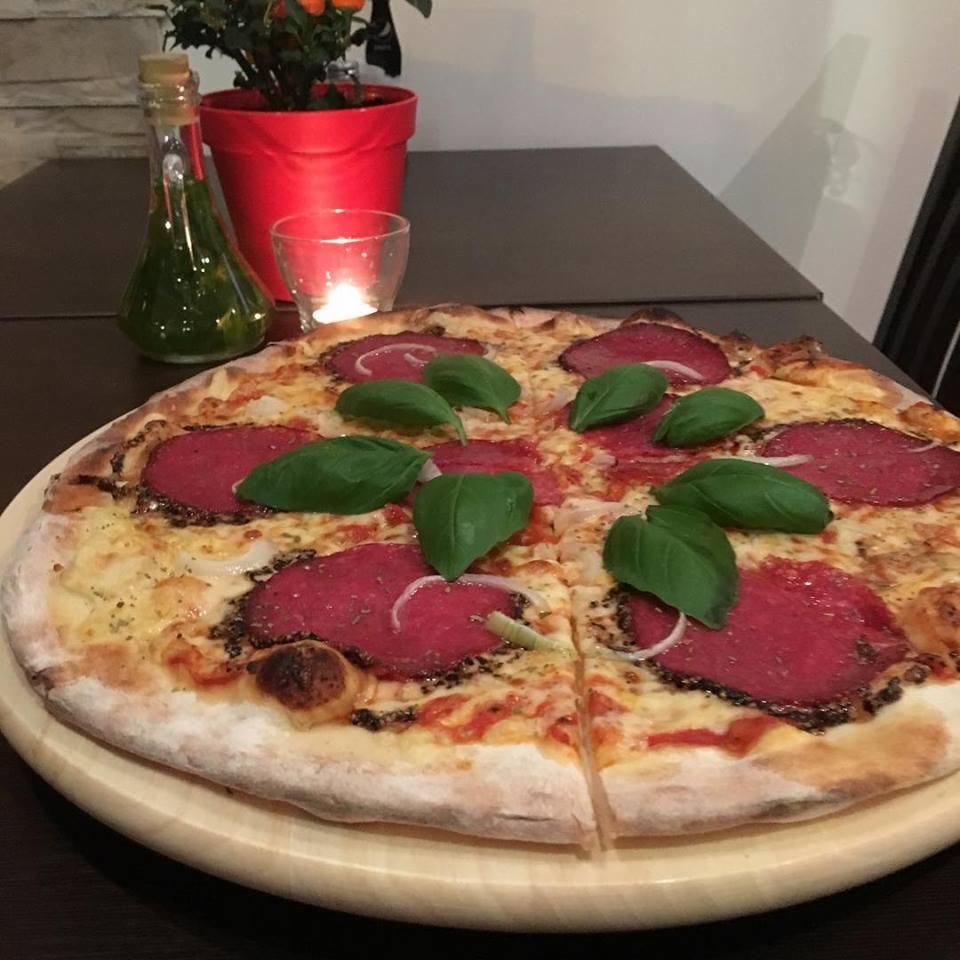 Pizza z salami, bazylią i cebulą na drewnianej desce, obok butelka z zielonym sosem, świeca i roślina w czerwonej doniczce.