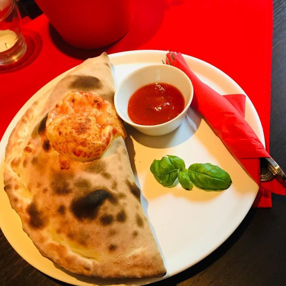 Calzone z dodatkiem sera i sosem pomidorowym w białej miseczce, udekorowane listkami bazylii, na czerwonym obrusie.