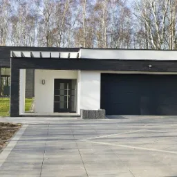 Nowoczesny dom z antracytową bramą garażową segmentową, widok z zewnątrz na podjazd wyłożony szarą kostką brukową, minimalizm w architekturze.