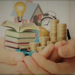 Dłonie trzymające stos książek z otwartą książką na wierzchu, model domu z żarówką, stosy monet, globus, zegar i model samochodu.