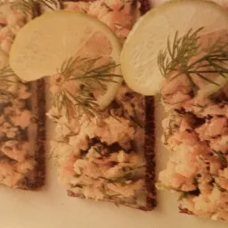 Kanapki z pastą rybną na ciemnym pieczywie, udekorowane plasterkiem cytryny i gałązką koperku.