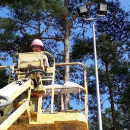 Pracownik w różowym kasku na podnośniku koszowym montuje reflektory na wysokim maszcie oświetleniowym na tle drzew