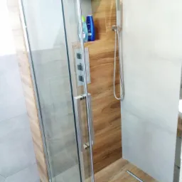 Nowoczesna kabina prysznicowa z drewnopodobnymi panelami, szklanymi drzwiami i chromowaną armaturą, uzupełniona o wnękę z kosmetykami Nivea.