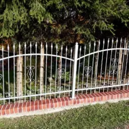 Białe, metalowe ogrodzenie z dekoracyjnymi elementami w kształcie serc i grotów, osadzone na niskim murku z czerwonej cegły, na tle zielonych drzew.