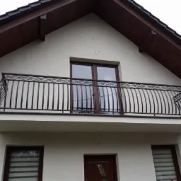 Czarna, metalowa balustrada balkonowa z dekoracyjnym wzorem na tle jasnej elewacji domu z brązowym dachem.