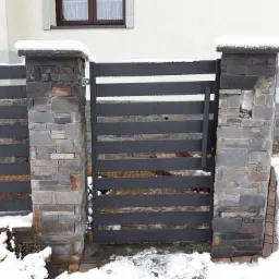 Nowoczesna brama posesyjna z poziomych, szarych profili, osadzona na kamiennych słupkach pokrytych śniegiem, z widocznym fragmentem elewacji budynku w tle.