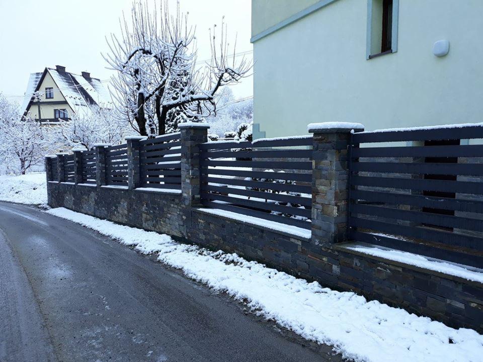 Nowoczesne ogrodzenie posesji z poziomych, grafitowych paneli na kamiennej podmurówce w zimowej scenerii, z domem w tle.