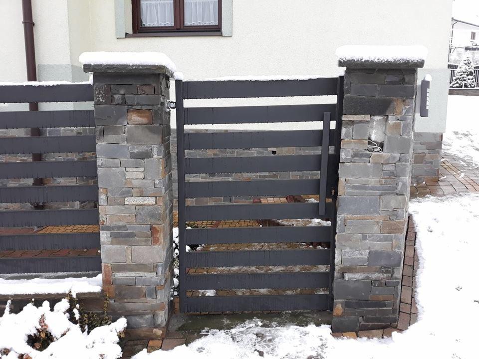Nowoczesna brama posesyjna z poziomych, szarych profili, osadzona na kamiennych słupkach pokrytych śniegiem, z widocznym fragmentem elewacji budynku w tle.