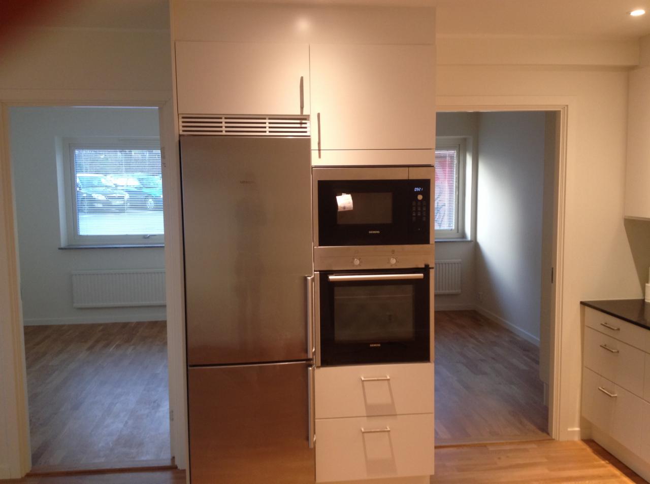 Nowoczesna kuchnia z zabudowaną lodówką ze stali nierdzewnej i sprzętem AGD marki Siemens, widok na sąsiedni pokój z drewnianą podłogą i oknem.