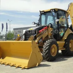 Żółta koparko-ładowarka Caterpillar 432E, widok z przodu, na placu z widocznymi flagami firmowymi w tle.