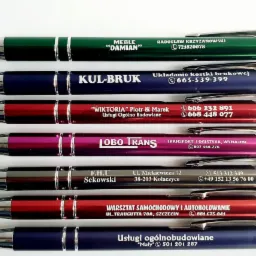 Kolekcja długopisów reklamowych w różnych kolorach z nadrukowanymi nazwami firm, adresami i numerami telefonów na białym tle.