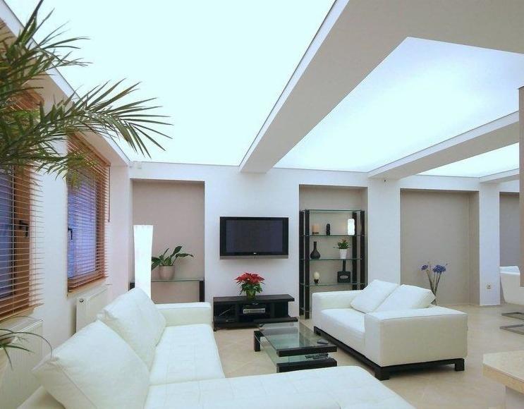 Nowoczesny salon z białymi sofami, przeszklonym stolikiem kawowym i podświetlanym sufitem napinanym z wbudowanym oświetleniem, widok na rośliny doniczkowe i telewizor na ścianie.