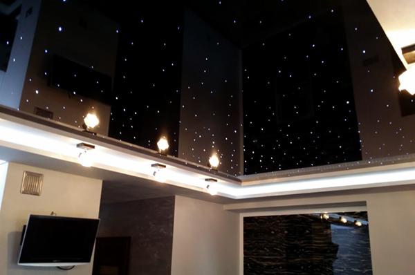 Czarny, błyszczący sufit z efektem gwiazd, oświetlony listwą LED i lampami punktowymi, widok na salon z szarą ścianą i telewizorem.