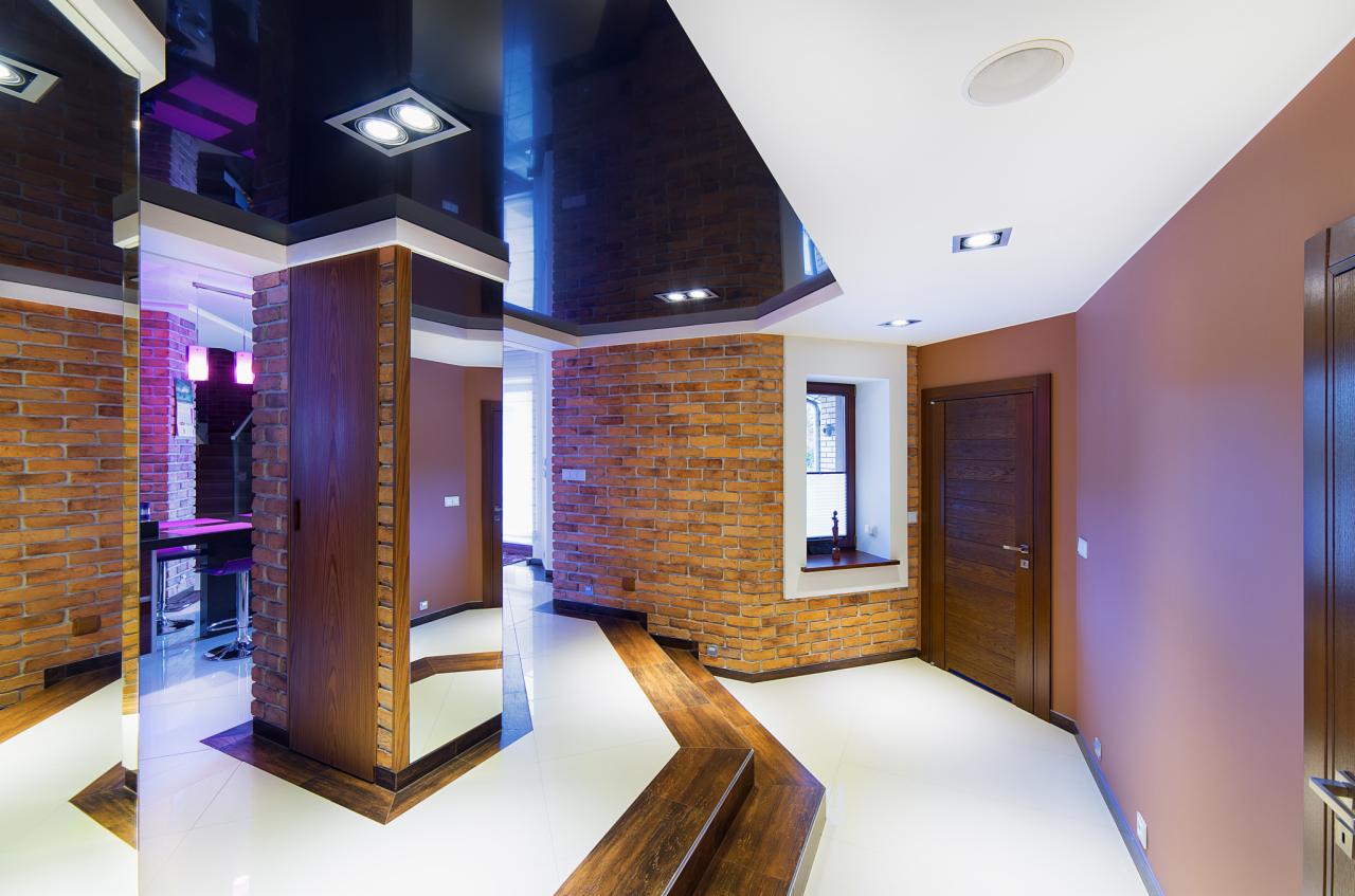 Nowoczesne wnętrze z ceglanymi ścianami, błyszczącym sufitem i nietypowym przejściem podłogowym z drewnianymi panelami, oświetlone punktowymi lampami.