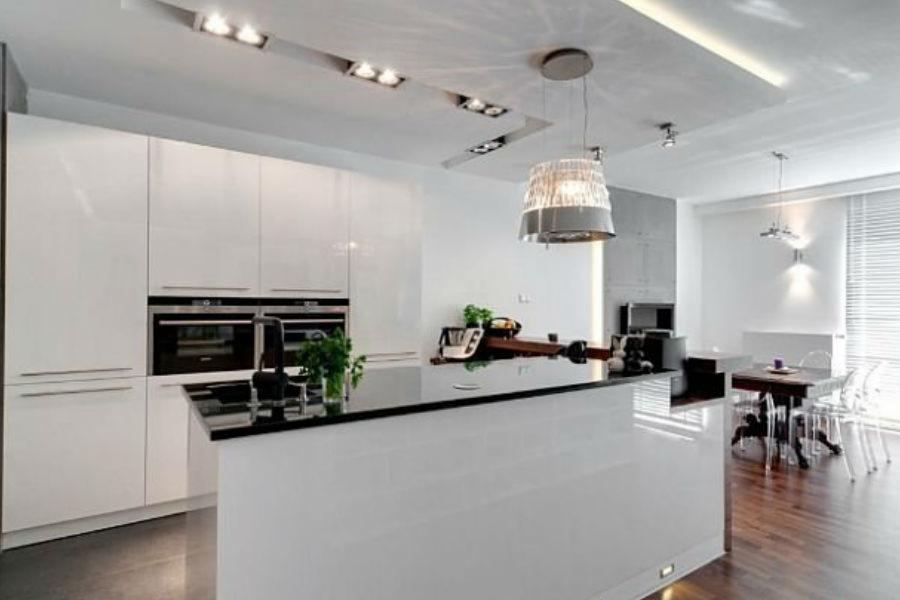 Nowoczesna kuchnia z białymi, lakierowanymi frontami, czarnym blatem i wyspą, oświetlona designerską lampą wiszącą i punktowymi reflektorami sufitowymi, z widoczną częścią jadalni z drewnianą podłogą.
