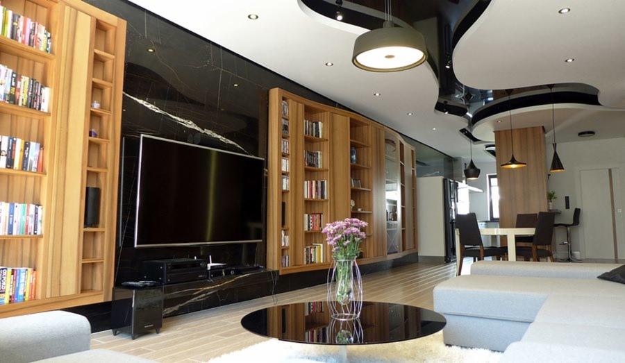 Elegancki salon z czarną marmurową ścianą, drewnianymi regałami na książki, dużym telewizorem i okrągłym stolikiem kawowym z bukietem kwiatów; sufit z podwieszanymi elementami i nowoczesnym...