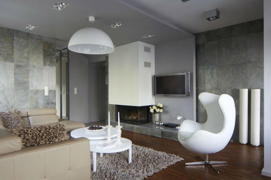 Elegancki salon z kamienną ścianą, nowoczesnym kominkiem, białym fotelem Egg i skórzaną sofą w odcieniach beżu, oświetlony punktowymi lampami sufitowymi i wiszącą lampą w kształcie półkuli.