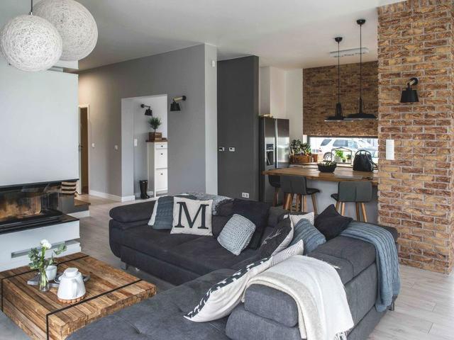 Nowoczesny salon z szarą sofą narożną, drewnianym stolikiem kawowym, ceglaną ścianą i otwartą kuchnią w tle, oświetlony wiszącymi lampami kulami.