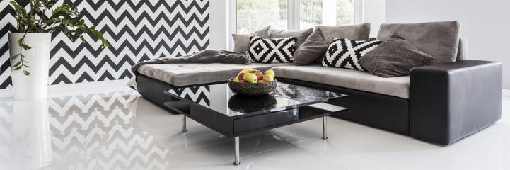 Nowoczesny salon z narożną sofą w odcieniach szarości, czarnym stolikiem kawowym z owocami w koszyku i białą, błyszczącą podłogą odbijającą wzór zygzakowatej tapety.