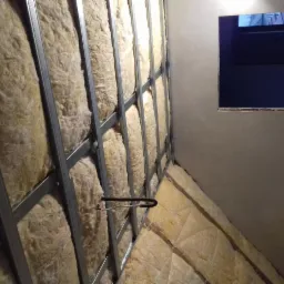 Ocieplenie poddasza wełną mineralną, widoczne metalowe profile konstrukcyjne, fragment okna dachowego i przewód elektryczny.