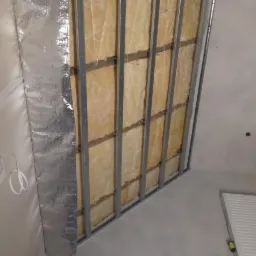 Wnętrze w trakcie prac wykończeniowych: widoczna konstrukcja metalowa, wypełniona wełną mineralną, przykryta folią aluminiową, przygotowana pod montaż płyt gipsowo-kartonowych.