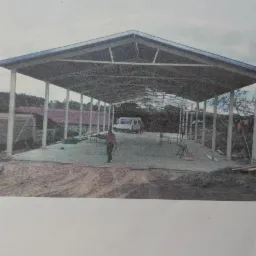 Wznoszona hala z konstrukcją stalową, widoczne słupy nośne, dach i częściowo betonowa podłoga, na zewnątrz zaparkowany biały bus.