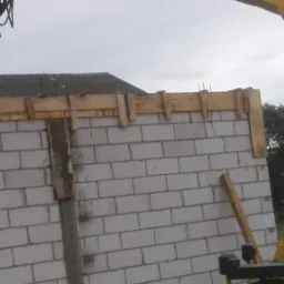 Wznoszenie ściany z bloczków betonowych z drewnianym szalunkiem górnym, widoczny fragment wysięgnika dźwigu teleskopowego.