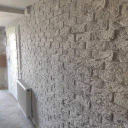 Ściana wykończona dekoracyjną cegłą w odcieniach szarości, z grzejnikiem przy ścianie i widocznym zabezpieczonym folią ościeżem drzwiowym w tle.